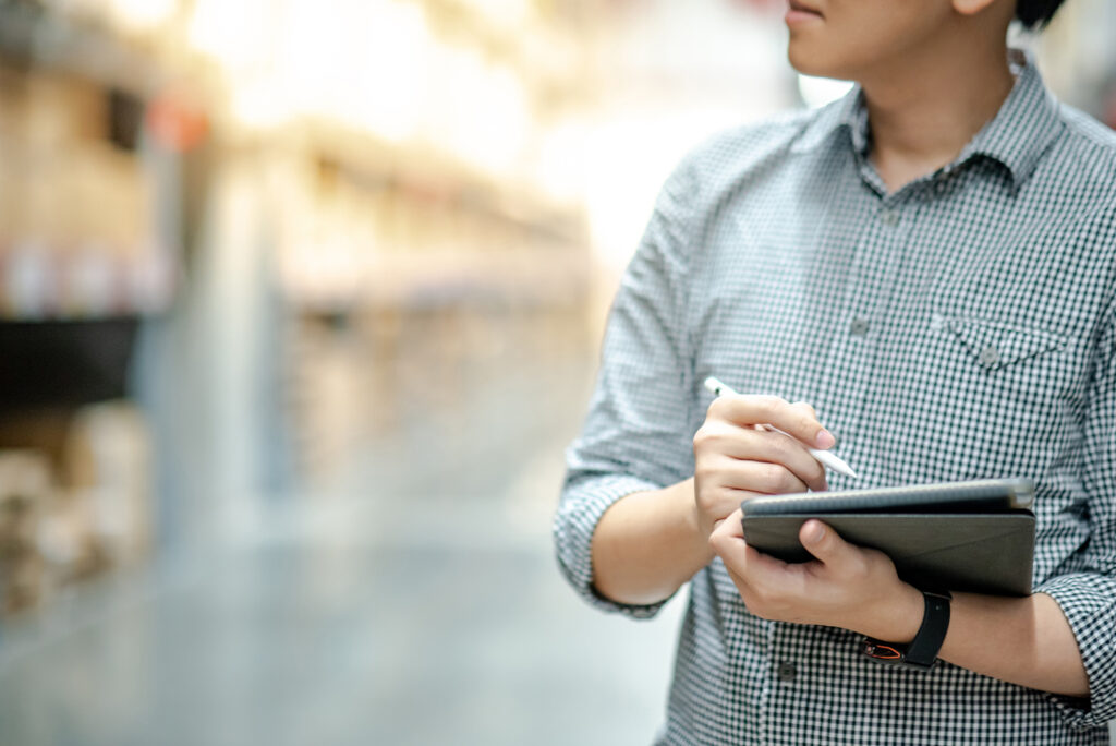 Young Asian Man Worker Doing Stocktaking Of Product In Cardboard Box On Shelves In Warehouse By Using Digital Tablet And Pen.