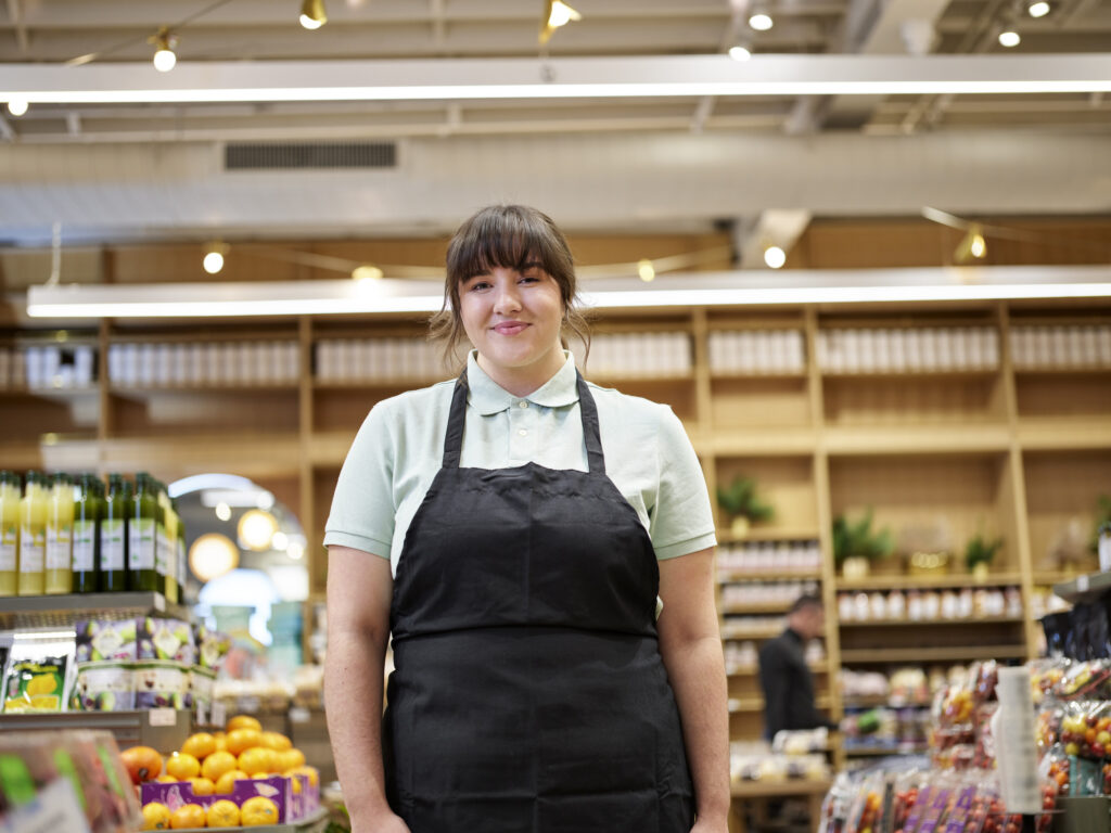 A female associate stands in a grocery store, smiling.