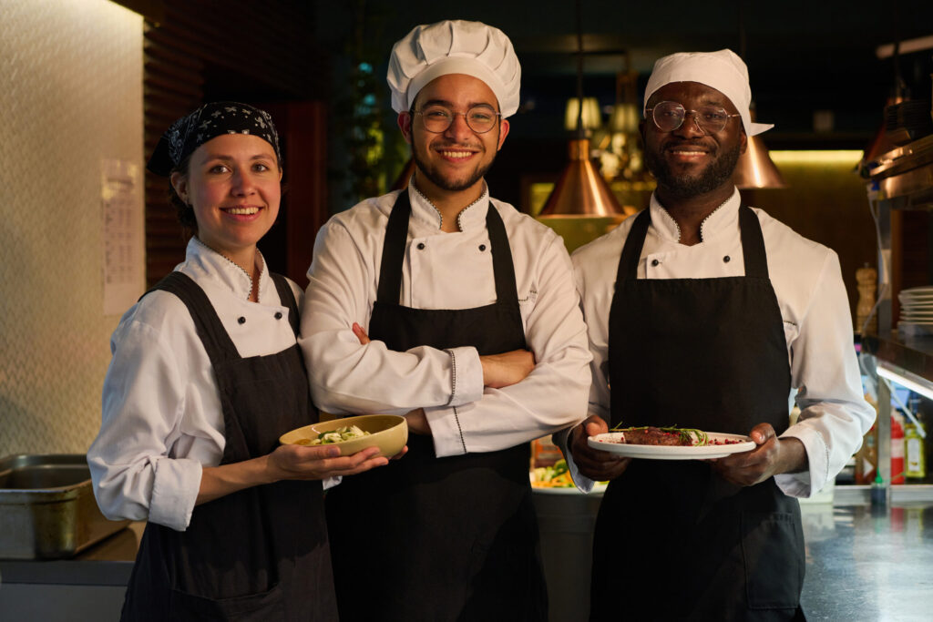 Group Of Young Intercultural Successful Cooks In Uniform Looking At Camera