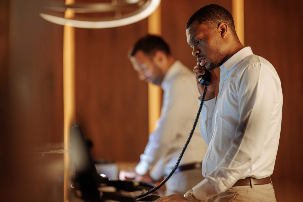 Two men in white shirts are working at desks; one is speaking on a corded phone while the other, a hospitality manager, is working on a computer. The background is blurred and has a warm tone.