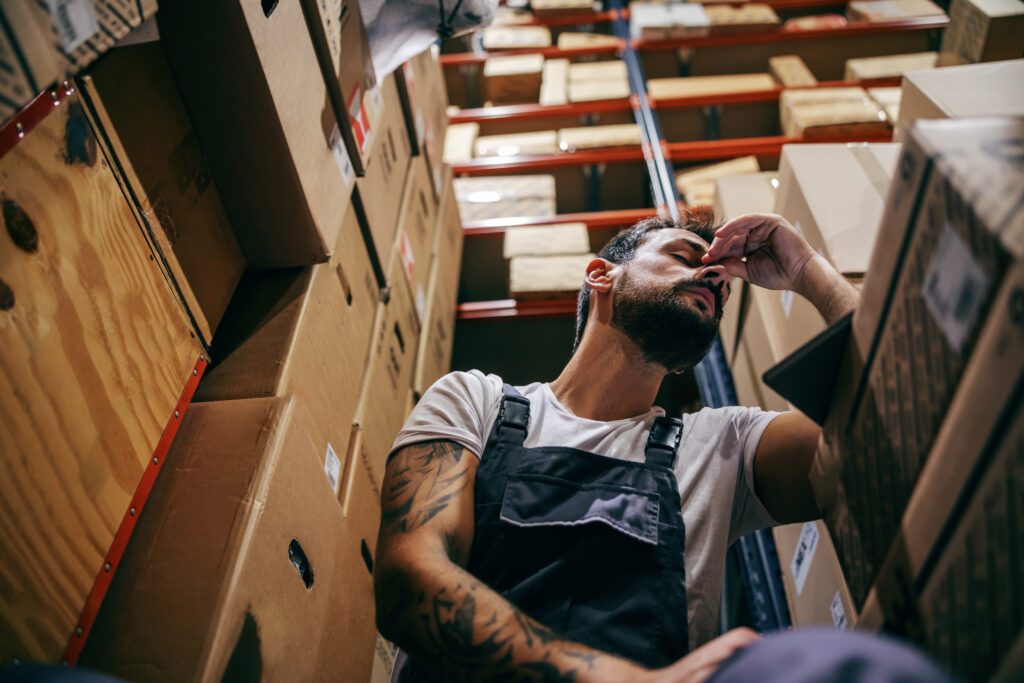 Tattooed Bearded Worker In Overalls Having Hard Day On The Work. Storage Of Import And Export Firm Interior.