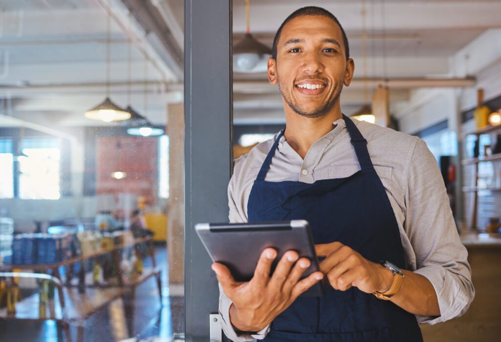 Restaurant Entrepreneur With Tablet, Leaning On Door And Open To Customers Portrait. Owner, Manager Or Employee Of A Startup Fast Food Store, Cafe Or Coffee Shop Business Standing Happy With A Smile