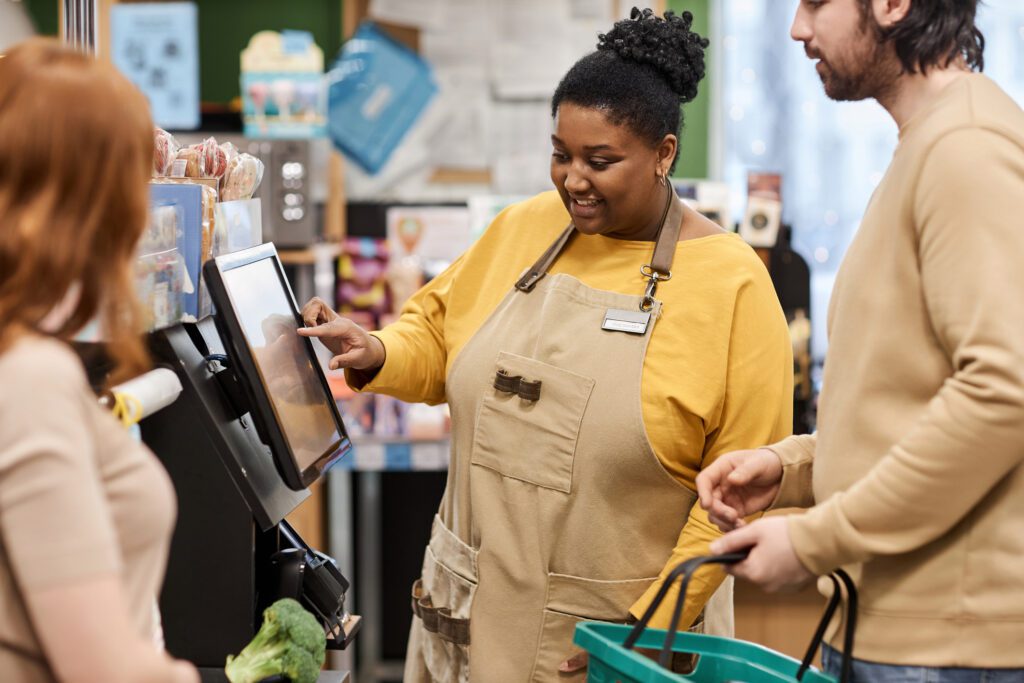 Smiling Black Woman Helping Customers With Self Checkout In Supermarket