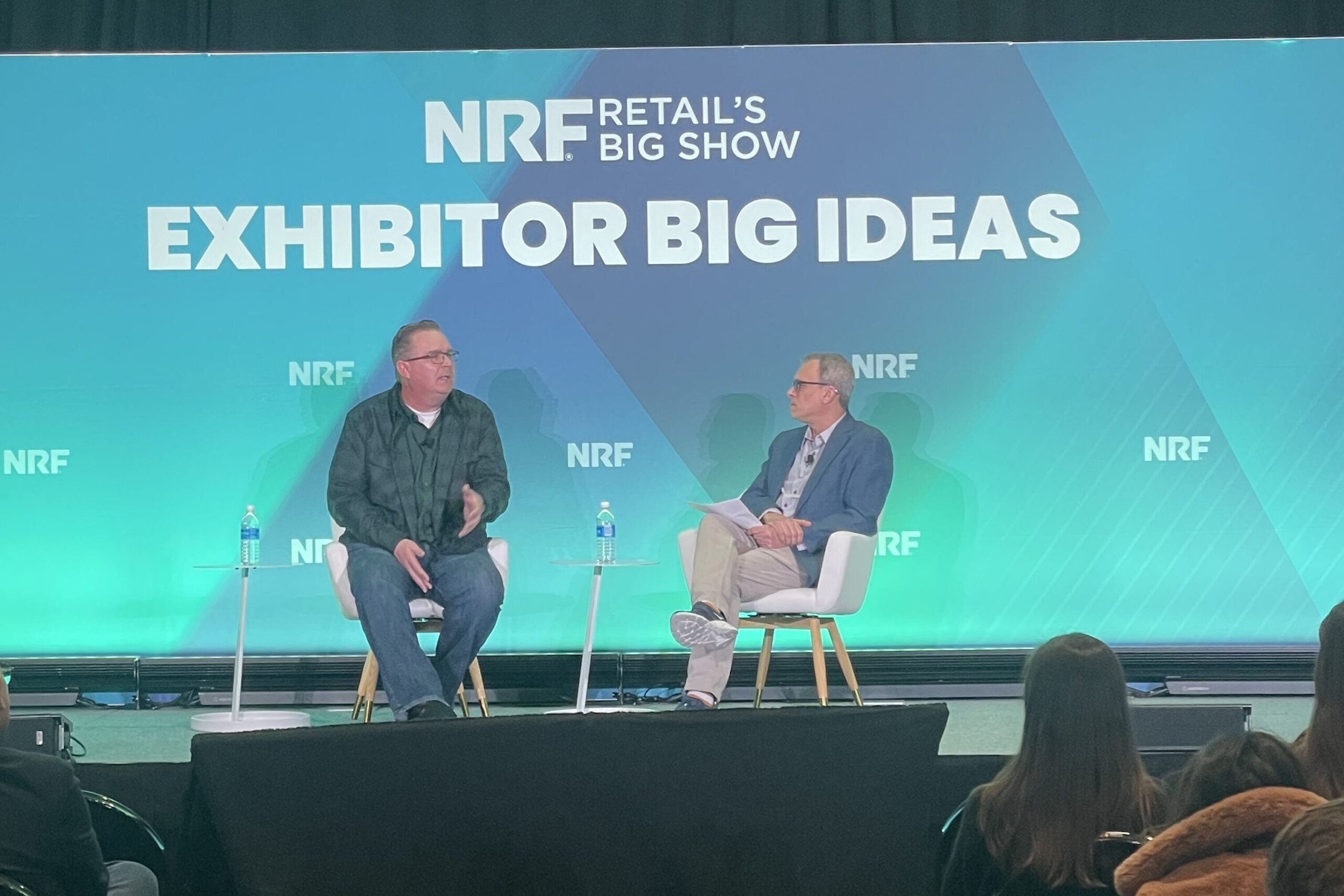 Image of an event stage where Kyle Eynon, Director of Training & Development at Sprouts Farmers Market is in conversation with Glenn Haussmann, hospitality expert and session host.
