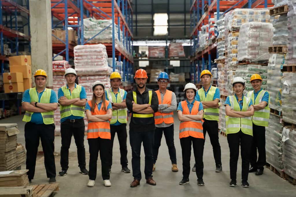 A diverse group of ten warehouse workers wearing safety vests and hard hats, standing with arms crossed in a large distribution center with high shelving.