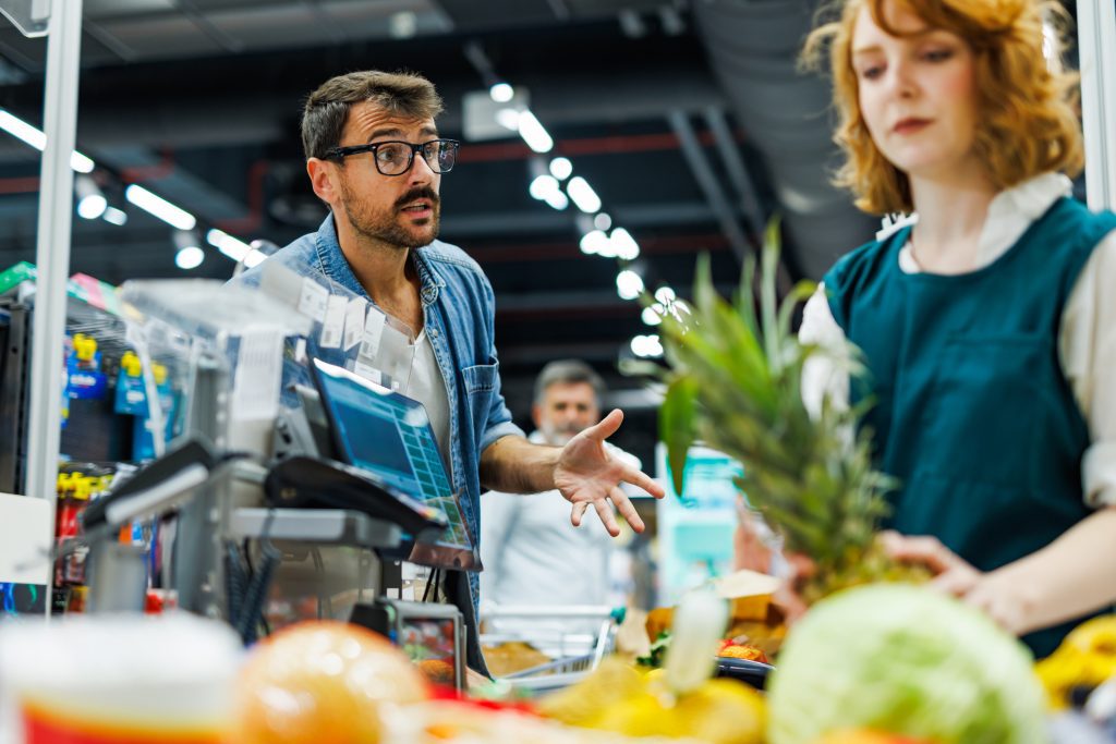 A retail scene showing a male customer in glasses looking frustrated and gesturing with his hand while speaking to a female cashier at a grocery checkout counter.