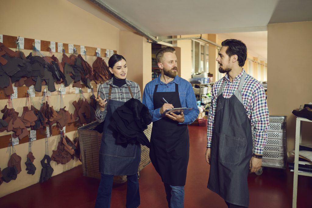 A scene inside a textile workshop where a female worker gestures toward hanging fabric patterns while discussing work with a male manager writing on a clipboard and a male colleague looking on.