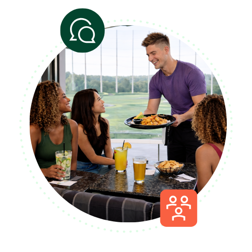 A male Topgolf server wearing a purple t-shirt smiles as he delivers a large platter of appetizers to three women seated at a table with cocktails and beers. The scene is accented by two floating circular icons: a dark green icon showing overlapping speech bubbles at the top left, and an orange icon depicting a team or group of people at the bottom right.