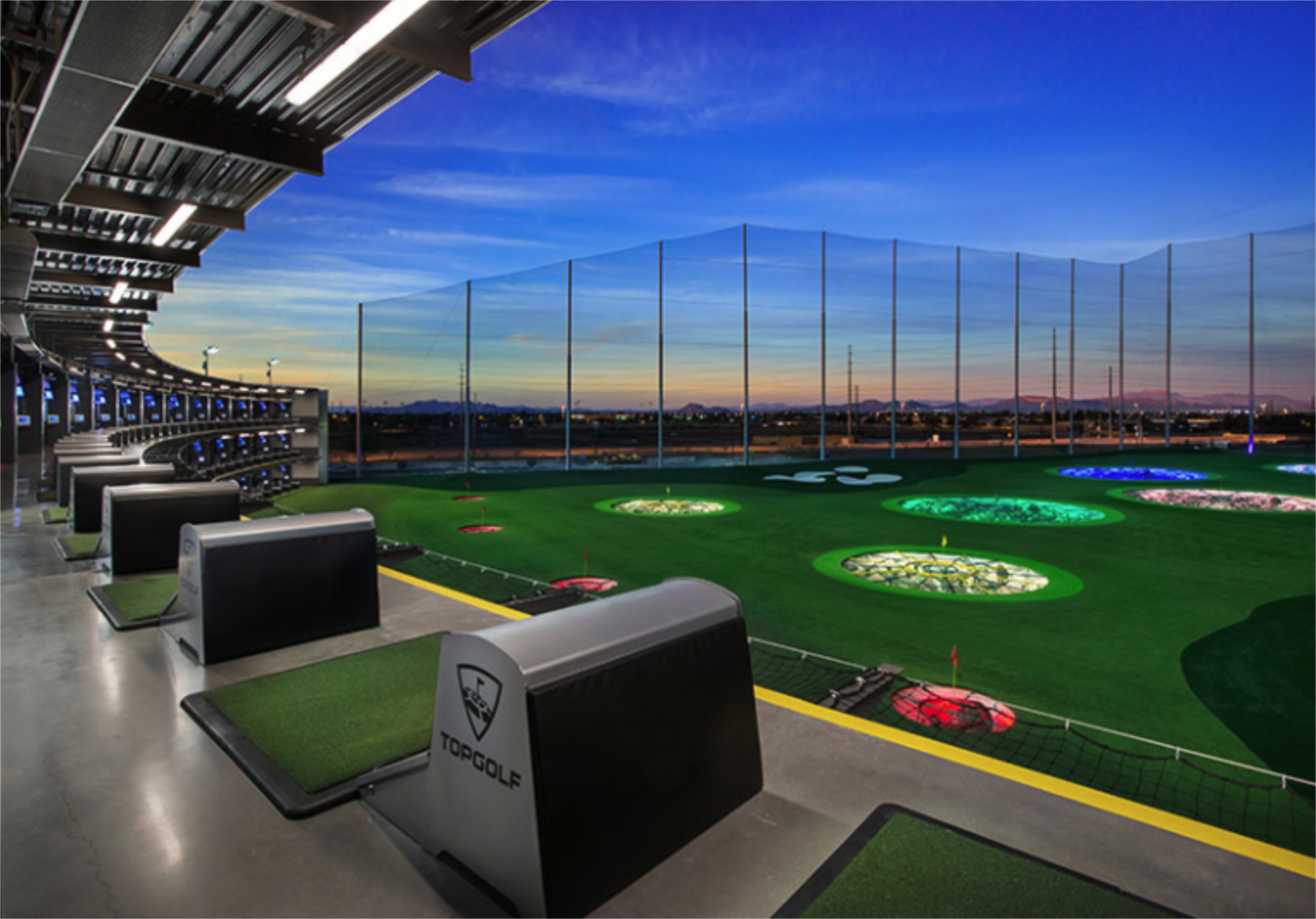 An outdoor driving range at a Topgolf facility showing a row of hitting bays with green turf mats under a covered structure. The green field features several large, illuminated circular target holes glowing in green, white, blue, and red under a twilight sky.