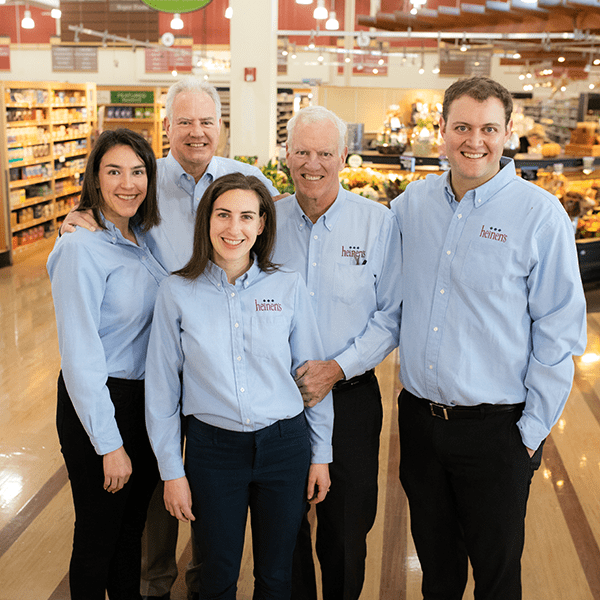 A group portrait of five smiling people posing together inside a Heinen's grocery store aisle. They are all wearing matching light blue button-down shirts, some embroidered with the Heinen's logo, paired with dark pants.