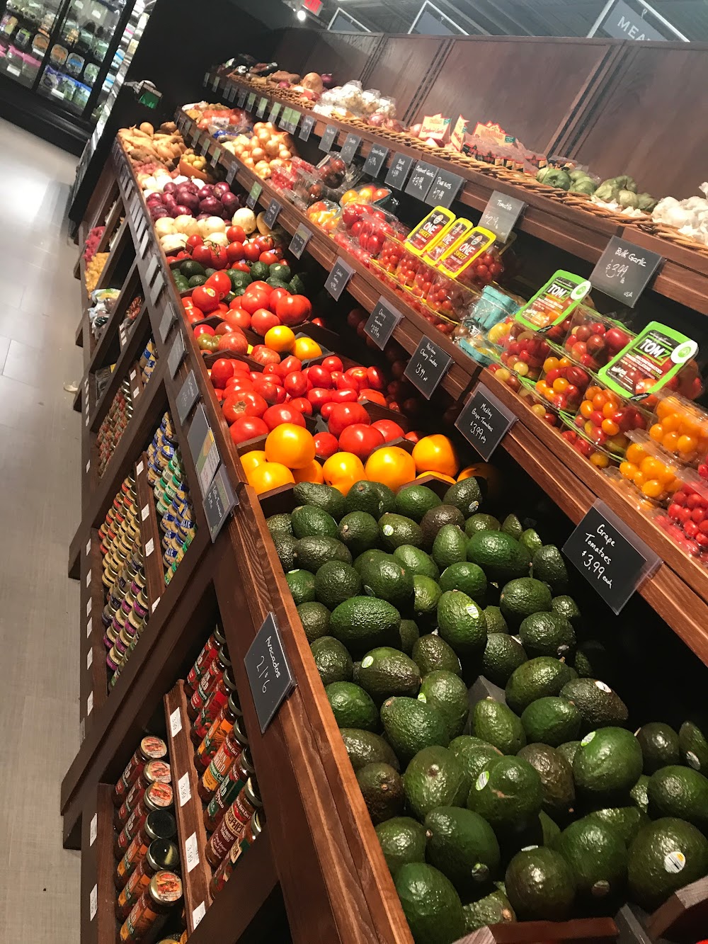 A high-angle view looking down a long, multi-tiered wooden produce display inside a grocery store. The shelves are filled with fresh items including an abundance of green avocados in the foreground, followed by orange citrus fruits, red tomatoes, small packaged grape tomatoes, onions, and various canned items stacked along the side bins.